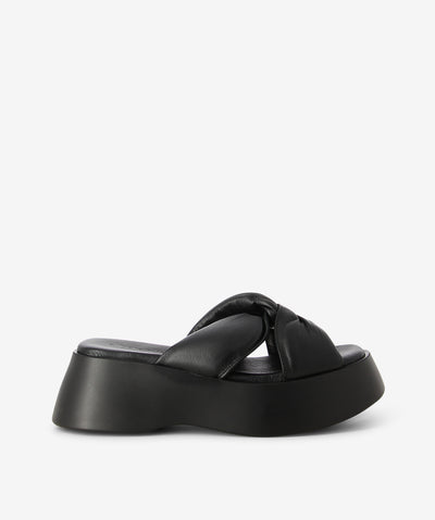 Black leather slides by 2 Baia Vista. It is a slip-on style that features two knotted crossover straps, a platform sole, and a rounded toe. 