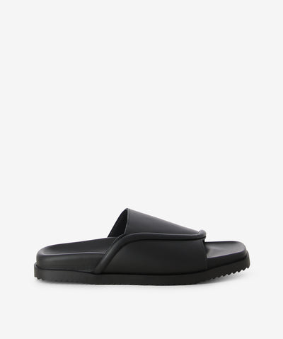 Black leather slides by Beau Coops. It is a slip-on style that features an oversized piped trim with a single leather strap, a moulded footbed, and a soft square toe. 